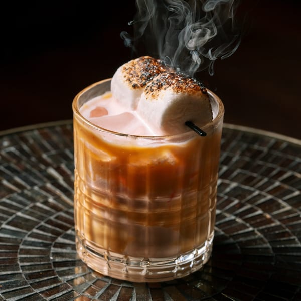 Smoked cocktail with toasted marshmallows and caramel drizzle in a glass on a dark, textured table.