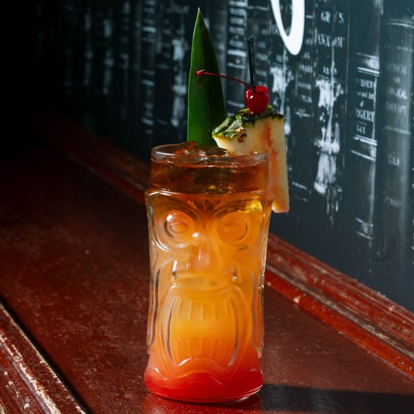 A colorful cocktail in a tiki glass, garnished with pineapple, a cherry, and a leaf.