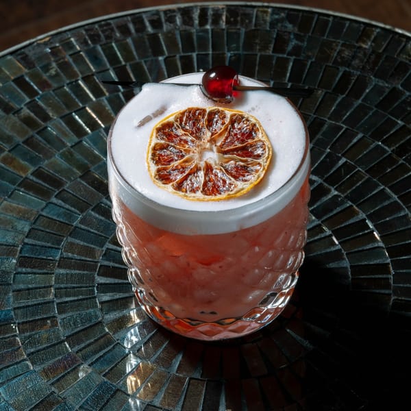 A foamy pink cocktail garnished with a dried orange slice and a cherry on a dark mosaic table.