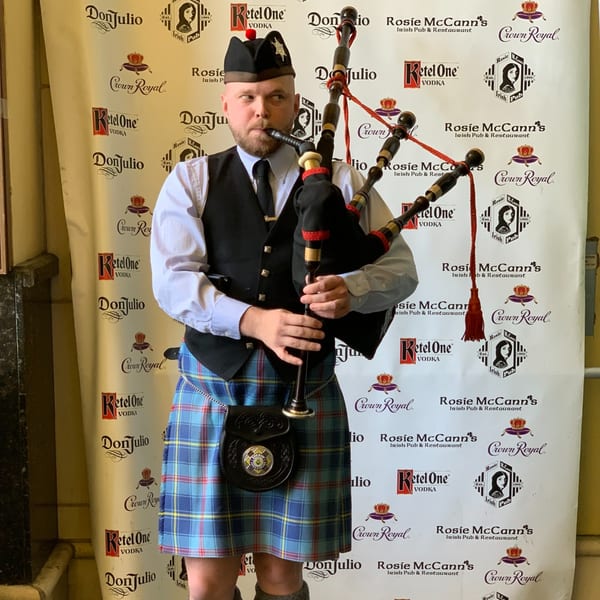 A bagpiper in a kilt plays against a Rosie McCann's Irish Pub & Restaurant branded backdrop.